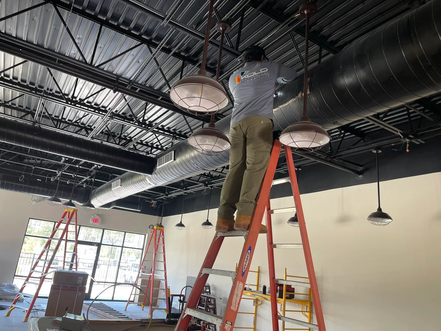 KOLD technician working from a ladder beneath exposed commercial ductwork.